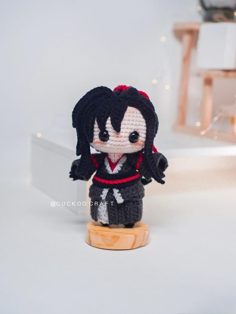 May include: A crocheted doll with black hair and a black and red outfit, standing on a small wooden base. The doll has a light beige face with black eyes. The text "@CUCKOOCRAFT" is visible on the base. The doll is a handmade item.