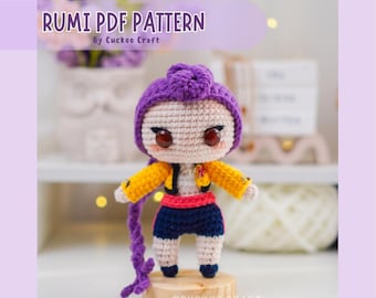 May include: A crocheted doll with a purple hat, yellow jacket, and blue shorts. The doll has large eyes and is standing on a wooden base. The text "RUMI PDF PATTERN" is displayed above the doll.