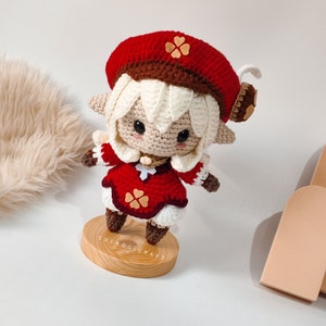 May include: A crocheted doll with white hair, a red beret with a brown trim and a flower, and a red dress with a white collar and brown boots. The doll is standing on a wooden base with the text "Cuckoo Craft".