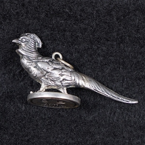Puede incluir: Un detallado dije de plata con forma de faisán, con un anillo para sujetarlo. El ave está de pie sobre una base circular. El dije tiene intrincados detalles de plumas y una larga cola. El fondo es una tela oscura y texturizada.
