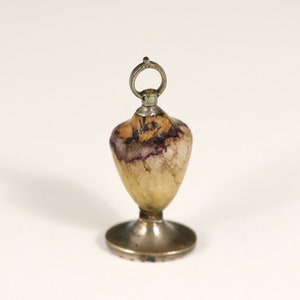 May include: A small, silver-colored bottle stopper with a heart-shaped, translucent, yellow and purple stone top. The stopper has a silver loop at the top for hanging.
