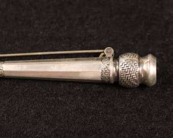 Puede incluir: Un broche plateado en forma de tubo largo y delgado con detalles intrincados. El broche tiene un pasador en un extremo y una tapa decorativa en el otro.