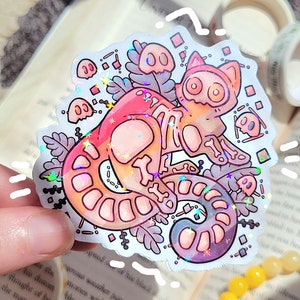 May include: A holographic sticker of a skeleton cat with a pink, orange, and purple gradient. The cat is surrounded by small ghosts and floral designs.