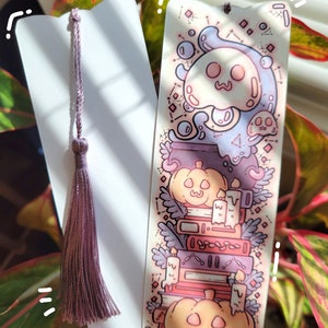 Two cute cardstock paper bookmarks with tassel and cat ear cut on the top held in a hand with a plant behind. One bookmark has an illustration of stacked books, pumpkins, ghosts, cauldron surrounded by plants and the other the white backside.