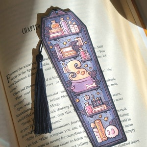 May include: A black and purple coffin-shaped bookmark with a black tassel. The bookmark features a shelf with various potions and books, including a cauldron with a skull and crossbones. The text "SPELL" is visible on the bookmark.