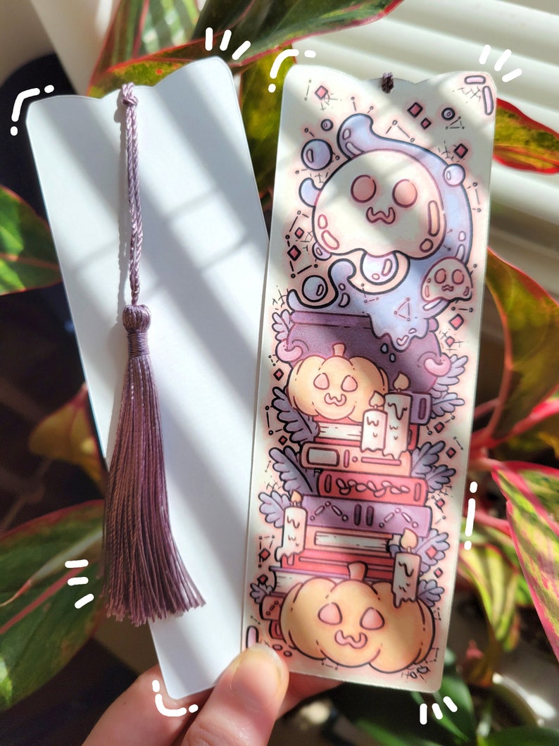 Two cute cardstock paper bookmarks with tassel and cat ear cut on the top held in a hand with a plant behind. One bookmark has an illustration of stacked books, pumpkins, ghosts, cauldron surrounded by plants and the other the white backside.