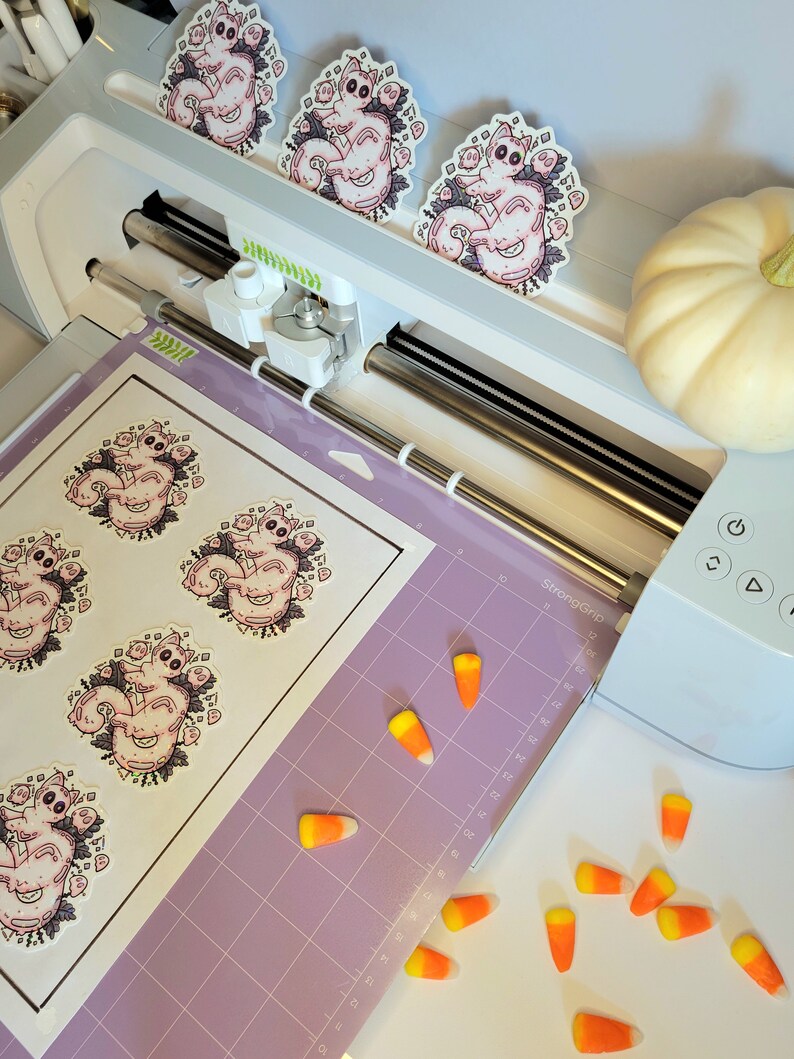 Holographic halloween ghost cat stickers. It is a light blue, white ghost surrounded by plant motifs and floating ghost friends. They are sitting on a purple Cricut mat ready to be cut in the manufacturing process surrounded by candy corn decorations