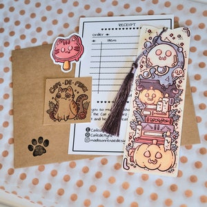 Cute cardstock paper bookmark with tassel and cat ear cut on the top resting on an packaging materials: receipt, sticker, tissue paper card. The bookmark has an illustration of stacked books, pumpkins, ghosts, and cauldron surrounded by plants.