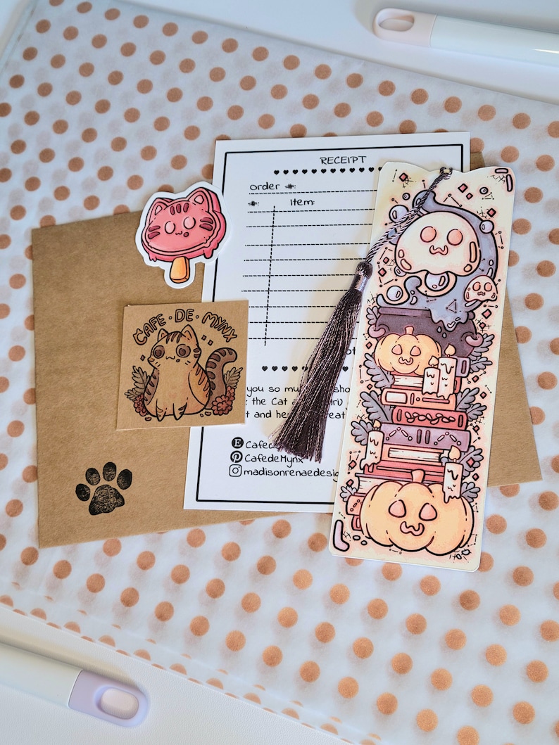 Cute cardstock paper bookmark with tassel and cat ear cut on the top resting on an packaging materials: receipt, sticker, tissue paper card. The bookmark has an illustration of stacked books, pumpkins, ghosts, and cauldron surrounded by plants.