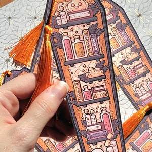 Gold orange coffin shaped bookcase bookmark with orange tassel. Features books, potions, pumpkin, candles and other witch library designs on the shelves.  One is held in a hand with more on a decorative white and gold background behind.