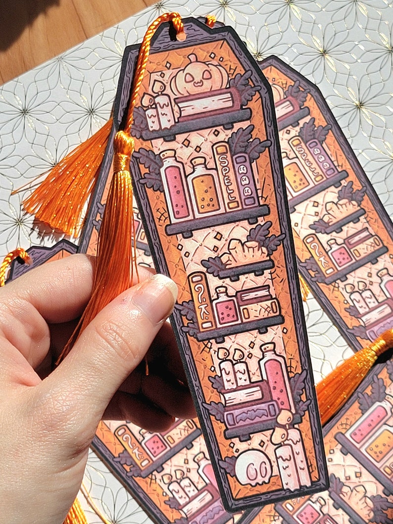 Gold orange coffin shaped bookcase bookmark with orange tassel. Features books, potions, pumpkin, candles and other witch library designs on the shelves.  One is held in a hand with more on a decorative white and gold background behind.