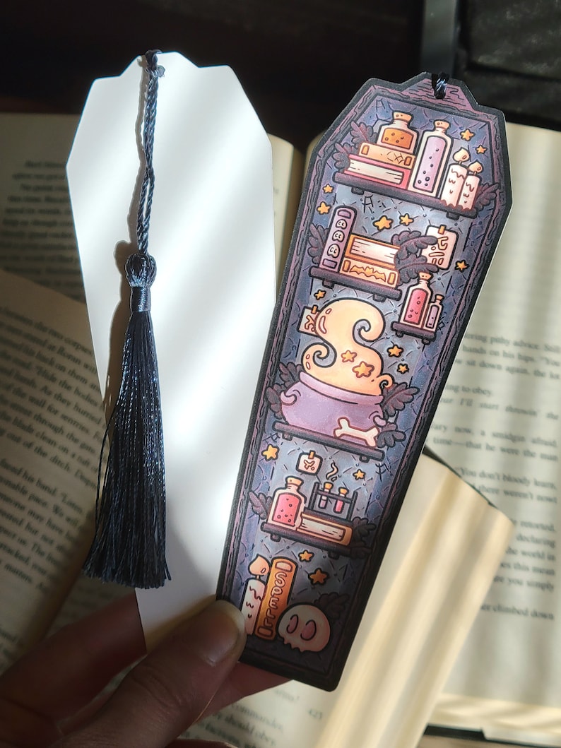 Navy blue coffin shaped bookcase bookmark with grey blue tassel. Features cauldron, skull, books, potions, and other witch library designs on the shelves. One shows the illustration on the front while another shows the white backside.