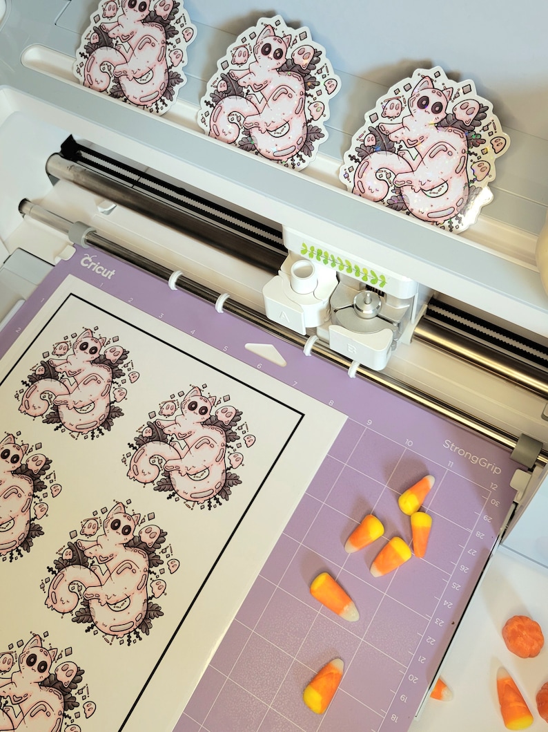 May include: A sheet of white sticker paper with six pink and white cat illustrations. The cat illustrations are surrounded by small white ghosts and black leaves. The paper is on a purple cutting mat with a Cricut cutting machine in the background.