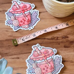 K&ouml;nnte beinhalten: Ein rosa-wei&szlig;er Aufkleber mit einer Cartoon-Illustration eines Boba-Tees mit einem Katzenpfoten-Deckel und einem Strohhalm. Der Boba-Tee ist von wei&szlig;en Blumen und gr&uuml;nen Bl&auml;ttern umgeben.