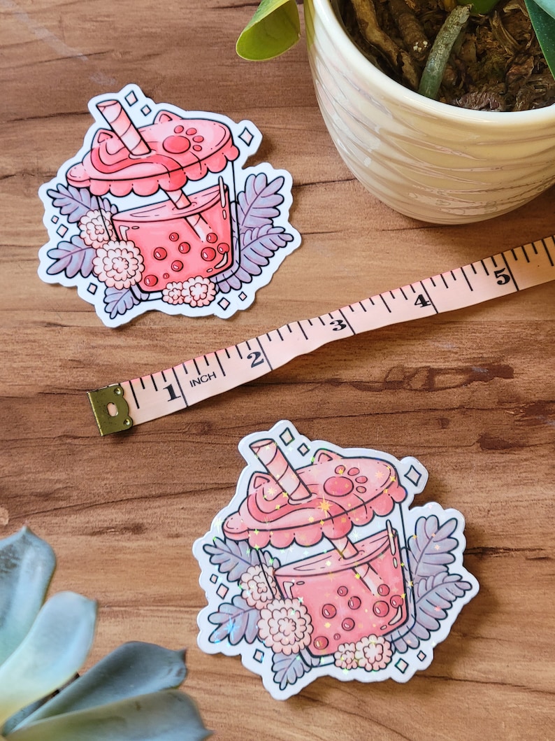 K&ouml;nnte beinhalten: Ein rosa-wei&szlig;er Aufkleber mit einer Cartoon-Illustration eines Boba-Tees mit einem Katzenpfoten-Deckel und einem Strohhalm. Der Boba-Tee ist von wei&szlig;en Blumen und gr&uuml;nen Bl&auml;ttern umgeben.
