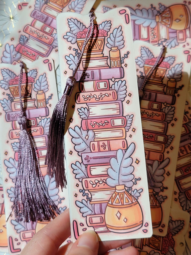 Cute cardstock paper bookmark with tassel and cat ear cut on the top held in hand. The bookmark has an illustration of aesthetic stacked pink and purple books surrounded by pretty plants. Behind are more bookmarks laying on a scrapbook paper square.