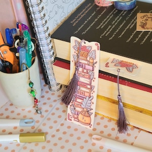 Cute cardstock paper bookmark with tassel and cat ear cut on the top leaning on a stack of books. The bookmark has an illustration of aesthetic stacked pink and purple books surrounded by pretty plants. It is surrounded by other books, pens, etc.