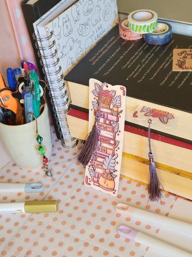 Cute cardstock paper bookmark with tassel and cat ear cut on the top leaning on a stack of books. The bookmark has an illustration of aesthetic stacked pink and purple books surrounded by pretty plants. It is surrounded by other books, pens, etc.