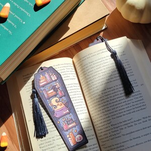 Navy blue coffin shaped bookcase bookmark with grey blue tassel. Features cauldron, skull, books, potions, and other witch library designs on the shelves. One sits on an open book while another is between the pages. The book rests on a wooden floor.