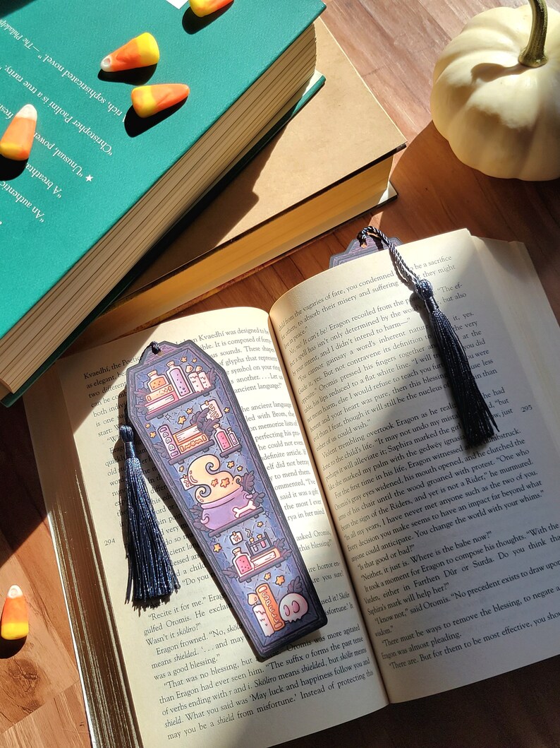 Navy blue coffin shaped bookcase bookmark with grey blue tassel. Features cauldron, skull, books, potions, and other witch library designs on the shelves. One sits on an open book while another is between the pages. The book rests on a wooden floor.