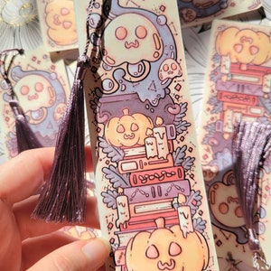 Cute cardstock paper bookmark with tassel and cat ear cut on the top held in hand. The bookmark has an illustration of stacked books, pumpkins, ghosts, and cauldron surrounded by plants. Behind are more bookmarks laying on a scrapbook paper.