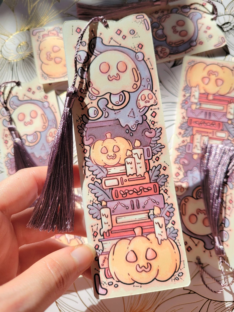 Cute cardstock paper bookmark with tassel and cat ear cut on the top held in hand. The bookmark has an illustration of stacked books, pumpkins, ghosts, and cauldron surrounded by plants. Behind are more bookmarks laying on a scrapbook paper.