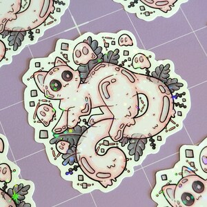 Holographic halloween ghost cat stickers. It is a light blue, white ghost surrounded by plant motifs and floating ghost friends. They are laying on a purple mat with a little bit of distance between them.