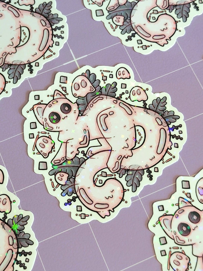 Holographic halloween ghost cat stickers. It is a light blue, white ghost surrounded by plant motifs and floating ghost friends. They are laying on a purple mat with a little bit of distance between them.