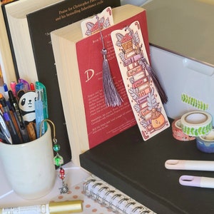 Cute cardstock paper bookmark with tassel and cat ear cut on the top leaning against a red book. The bookmark has an illustration of aesthetic stacked pink and purple books surrounded by pretty plants. It is surrounded by other books, pens, etc.