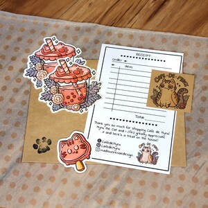 K&ouml;nnte beinhalten: Ein brauner Papierumschlag mit einem Pfotenabdruck-Aufkleber. Eine Quittung mit dem Text "Cafe de Mynx" und einer Comic-Katze mit einem Boba-Tee. Es gibt drei Aufkleber mit Boba-Tee mit einer Comic-Katze auf jedem.