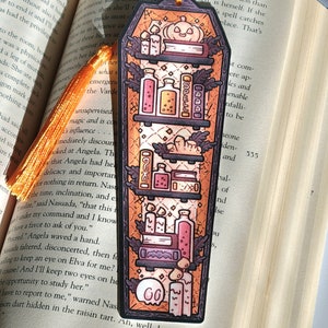 Gold orange coffin shaped bookcase bookmark with orange tassel. Features books, potions, pumpkin, candles and other witch library designs on the shelves.  It rests on an open book.