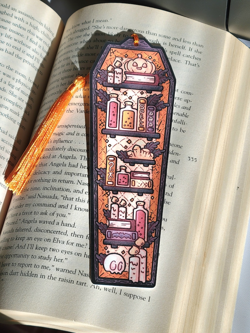 Gold orange coffin shaped bookcase bookmark with orange tassel. Features books, potions, pumpkin, candles and other witch library designs on the shelves.  It rests on an open book.