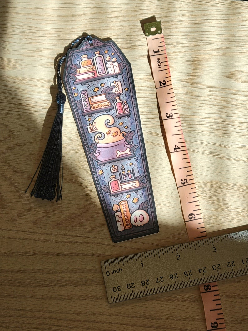 Navy blue coffin shaped bookcase bookmark with grey blue tassel. Features cauldron, skull, books, potions, and other witch library designs on the shelves. It rests on a light wood table with rulers beside to show size (roughly 6 inches by 2 inches).