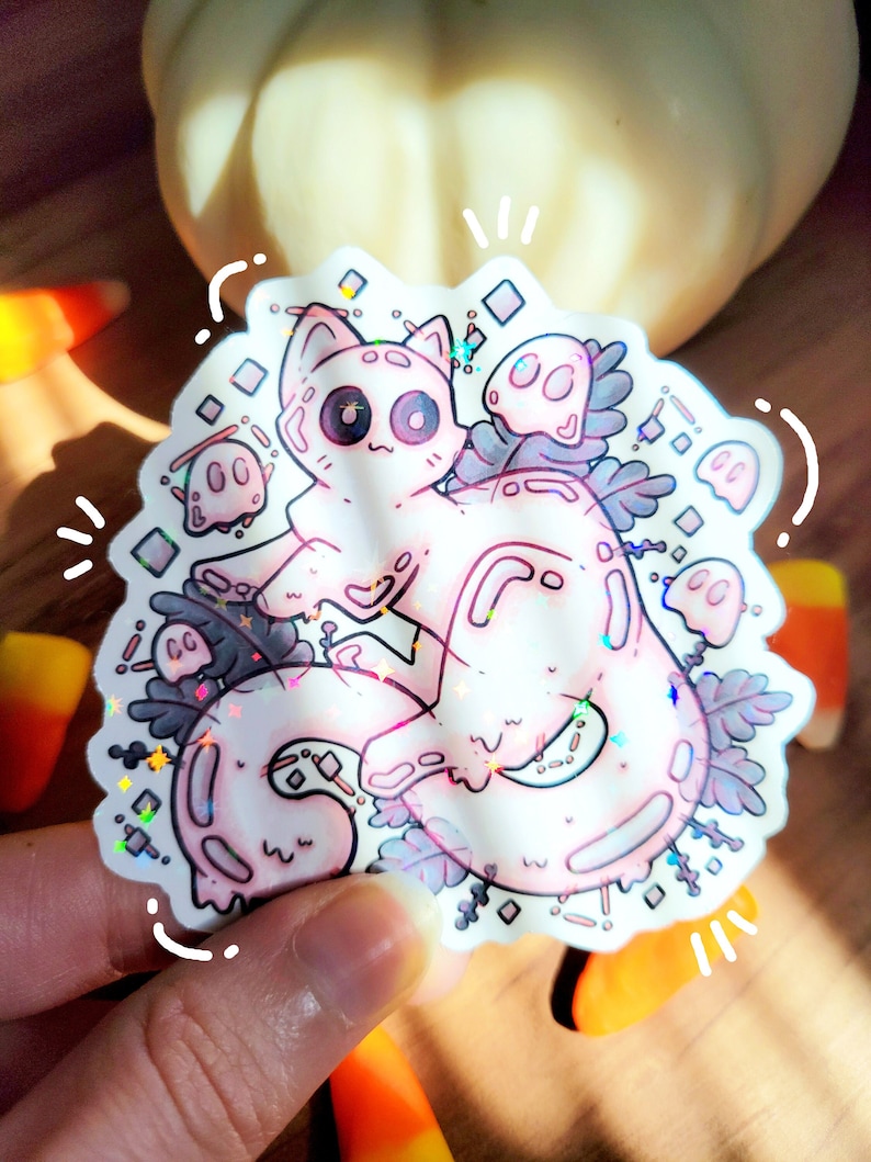 Holographic halloween ghost cat sticker. It is a light blue, white ghost surrounded by plant motifs and floating ghost friends. It is held in someones hand with pumpkin and candy corn decorations on a wood floor behind.