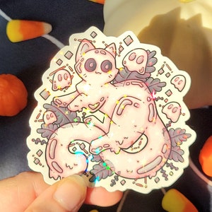 Holographic halloween ghost cat sticker. It is a light blue, white ghost surrounded by plant motifs and floating ghost friends. It is held in someones hand with a pumpkin and candy corn decorations behind.