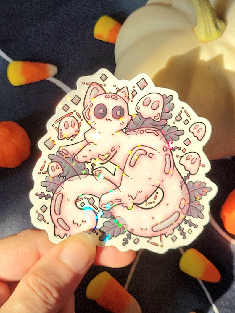 Holographic halloween ghost cat sticker. It is a light blue, white ghost surrounded by plant motifs and floating ghost friends. It is held in someones hand with a pumpkin and candy corn decorations behind.