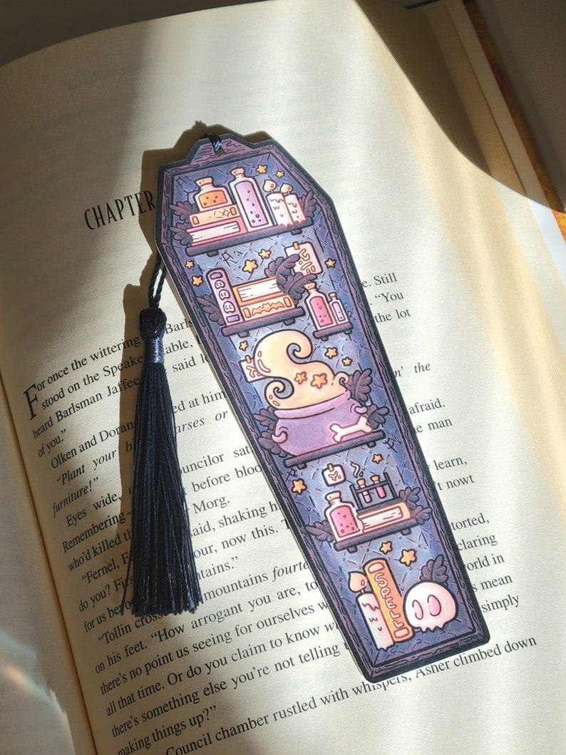 Navy blue coffin shaped bookcase bookmark with grey blue tassel. Features cauldron, skull, books, potions, and other witch library designs on the shelves. It sits on an open book.