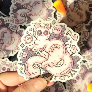 Holographic halloween ghost cat sticker. It is a light blue, white ghost surrounded by plant motifs and floating ghost friends. It is held in someones hand with more of the same sticker laying behind it.