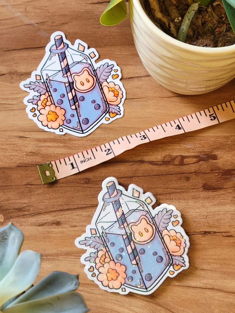 May include: A sticker featuring a cartoon illustration of a glass bottle with a straw, filled with a blue liquid and boba pearls. The bottle is surrounded by flowers and hearts. The sticker is on a wooden surface.