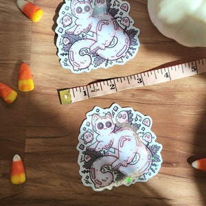 May include: A holographic sticker featuring a pink cat with a white face and large eyes surrounded by small ghosts and leaves. The sticker is on a wooden surface.