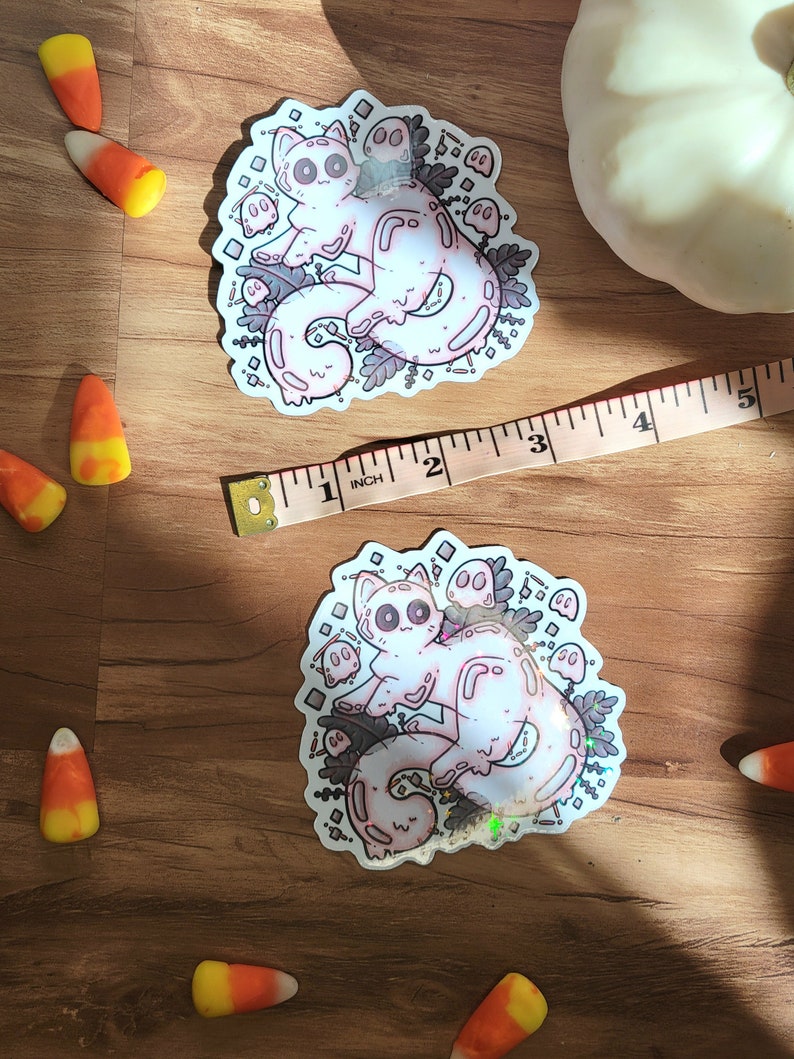 May include: A holographic sticker featuring a pink cat with a white face and large eyes surrounded by small ghosts and leaves. The sticker is on a wooden surface.