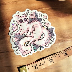 Holographic halloween ghost cat sticker. It is a light blue, white ghost surrounded by plant motifs and floating ghost friends. It is laying on a wooden floor with a pumpkin decoration and a pink ruler. The ruler measures 3in in size.