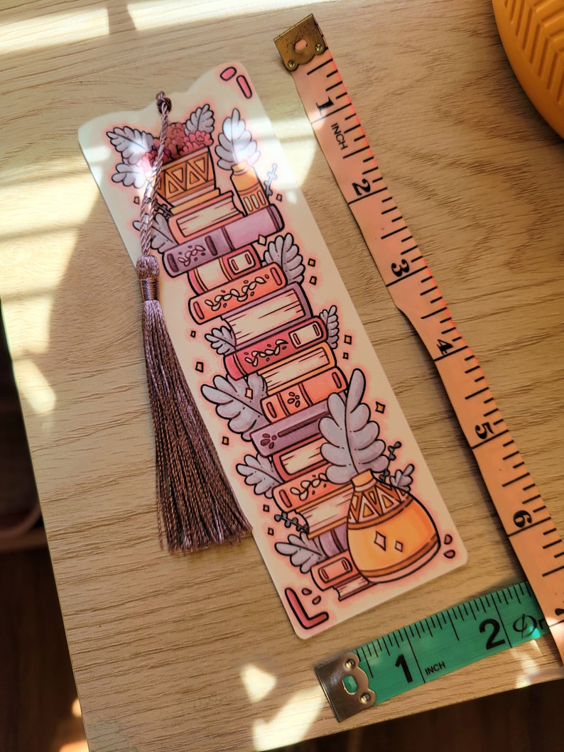 Cute cardstock paper bookmark with tassel and cat ear cut on top of a light wood table. The bookmark has an illustration of aesthetic stacked pink and purple books surrounded by pretty plants. Two tape measures sit beside the bookmark to show scale.