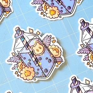 May include: A sticker featuring a cartoon illustration of a glass bottle filled with a blue drink with boba pearls and a straw. The bottle is decorated with flowers and hearts. The sticker is printed on white paper.