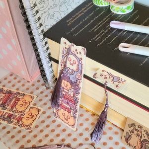 Cute cardstock paper bookmark with tassel and cat ear cut on the top leaning on a stack of books. The bookmark has an illustration of stacked books, pumpkins, ghosts, cauldron surrounded by plants. It is surrounded by other books, pens, etc