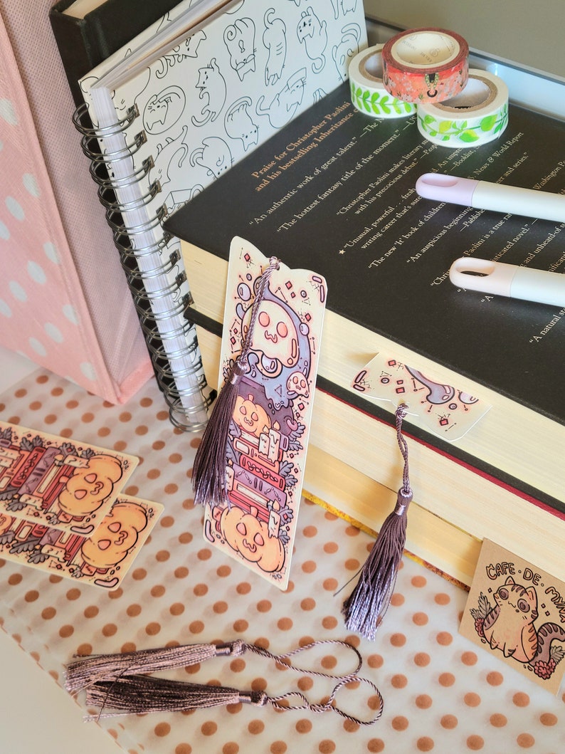 Cute cardstock paper bookmark with tassel and cat ear cut on the top leaning on a stack of books. The bookmark has an illustration of stacked books, pumpkins, ghosts, cauldron surrounded by plants. It is surrounded by other books, pens, etc