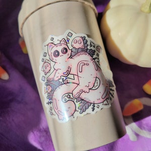 Holographic halloween ghost cat sticker. It is a light blue, white ghost surrounded by plant motifs and floating ghost friends. It is sitting on a drink bottle on purple cloth with a pumpkin and candy corn decorations surrounding it.