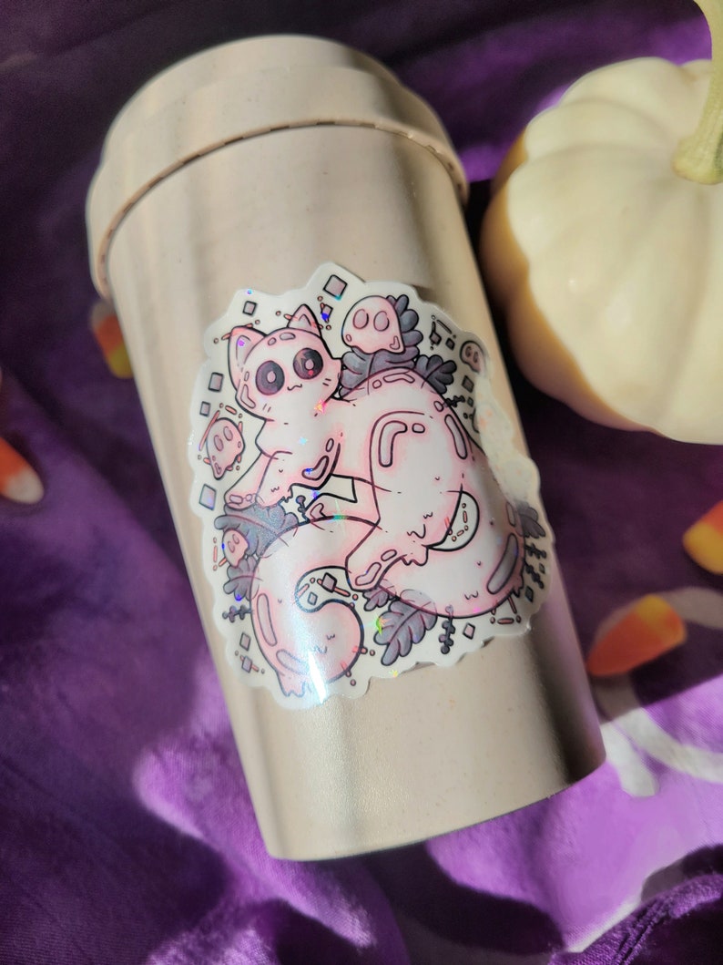 Holographic halloween ghost cat sticker. It is a light blue, white ghost surrounded by plant motifs and floating ghost friends. It is sitting on a drink bottle on purple cloth with a pumpkin and candy corn decorations surrounding it.