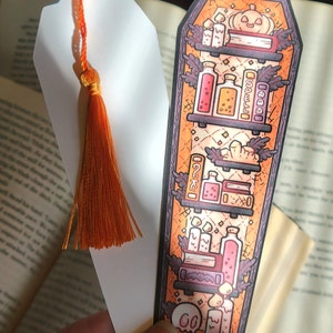 Gold orange coffin shaped bookcase bookmark with orange tassel. Features books, potions, pumpkin, candles and other witch library designs on the shelves.  One shows the illustrated front and another shows the white backside.