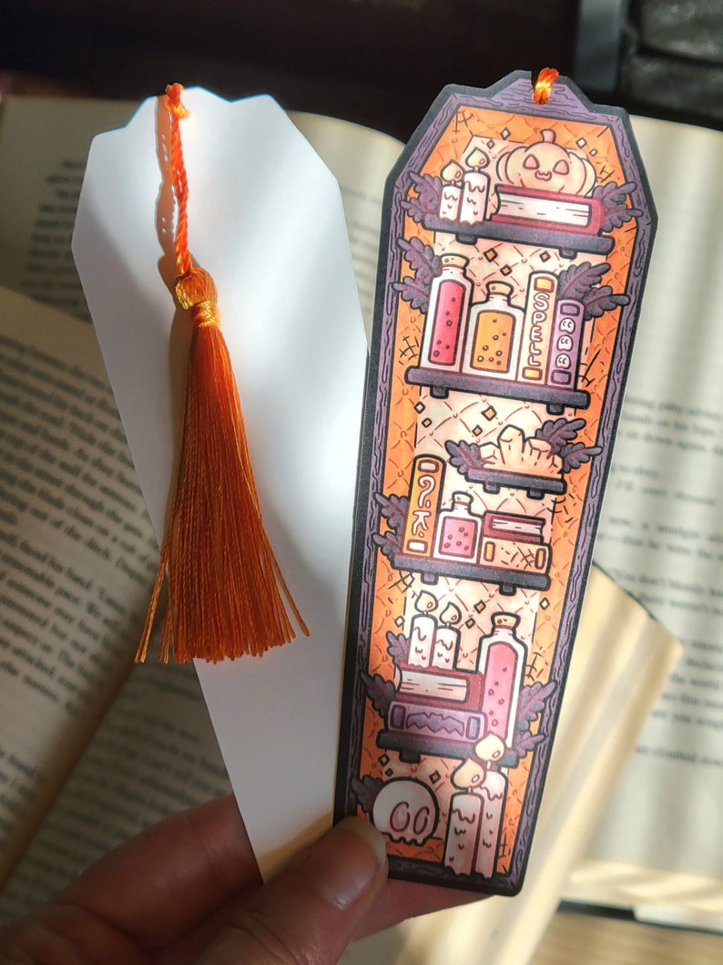 Gold orange coffin shaped bookcase bookmark with orange tassel. Features books, potions, pumpkin, candles and other witch library designs on the shelves.  One shows the illustrated front and another shows the white backside.